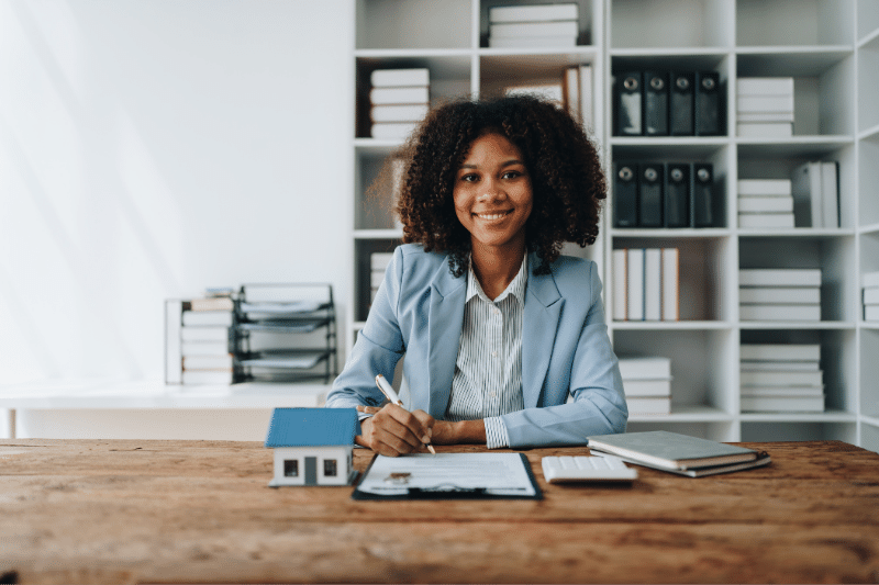 A woman in a light blue blazer sits at a desk, smiling, with documents, a pen, a small house model, a notebook, and a calculator in front of her. Bookshelves with files and books are in the background.